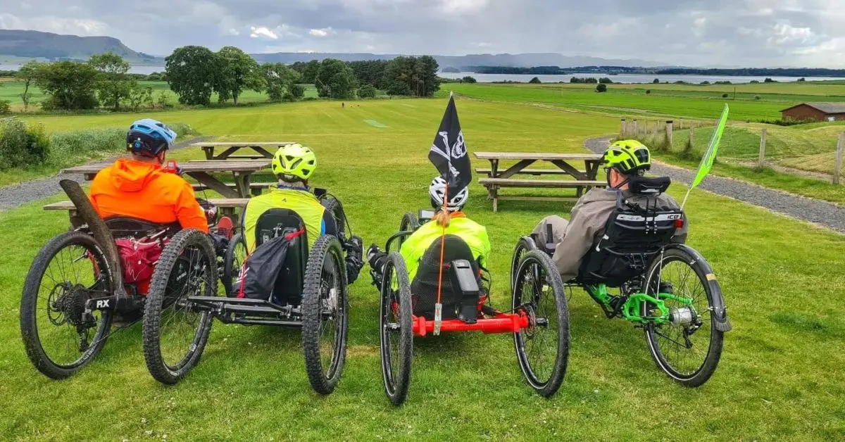 Four people riding adapted cycles across a grassy park beside water