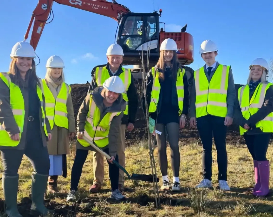 A group in hard hats and hi-vis vests at a groundbreaking, with a digger behind them