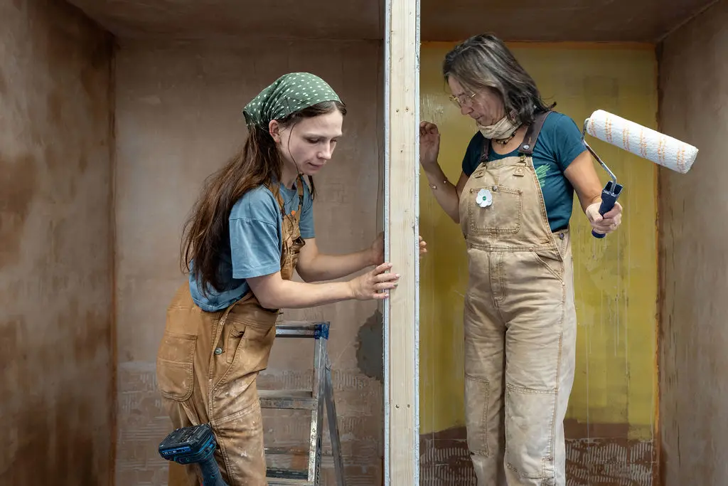 Two people in work dungarees decorating a room, one with a paint roller