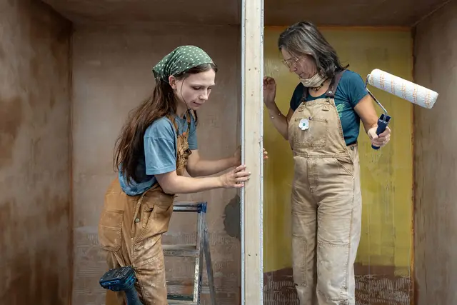 Two people in work dungarees decorating a room, one with a paint roller