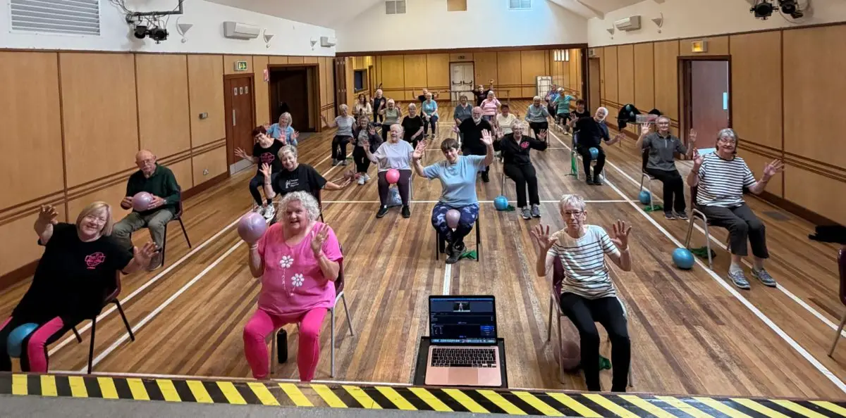 A seated exercise class in a community hall, with participants raising their arms