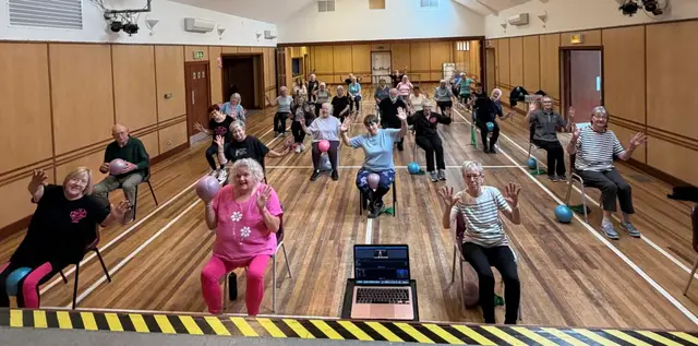 A seated exercise class in a community hall, with participants raising their arms