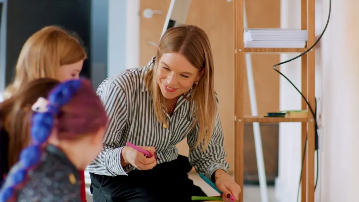 A smiling woman talks with a group of young people gathered round a table, one holding a pen